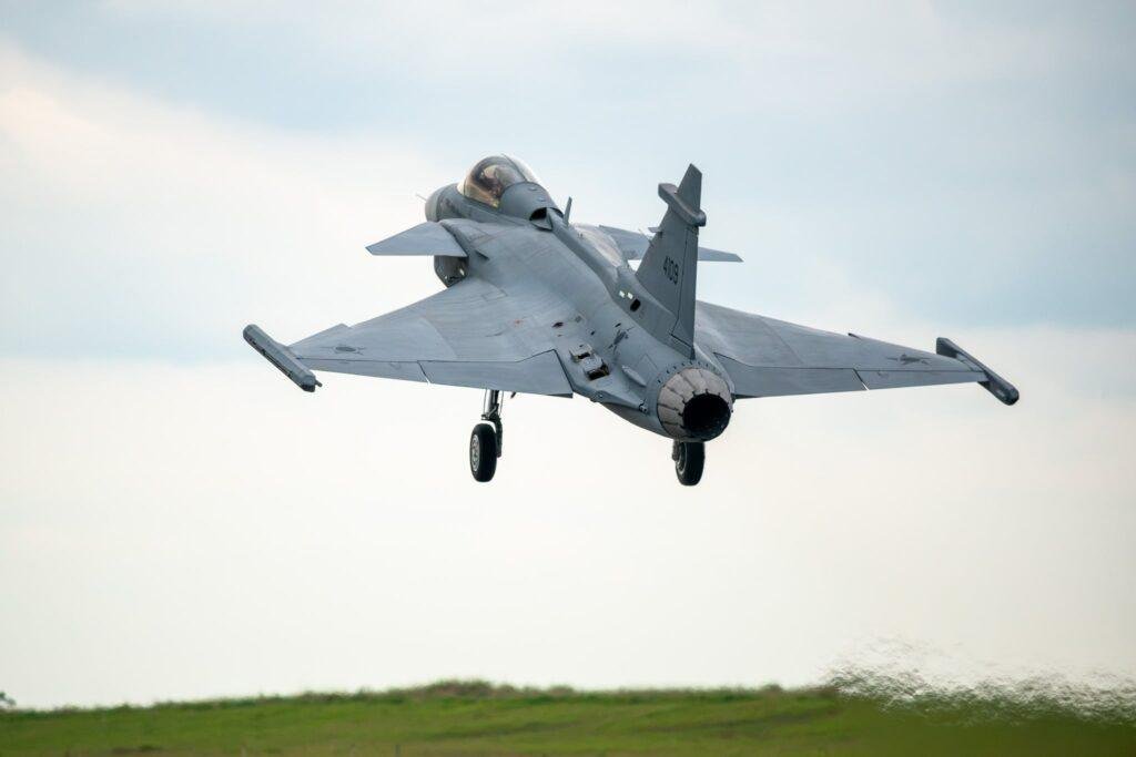 Brazil-built Gripen E fighter jet flying with landing gear extended during a low-altitude flight near an airfield.