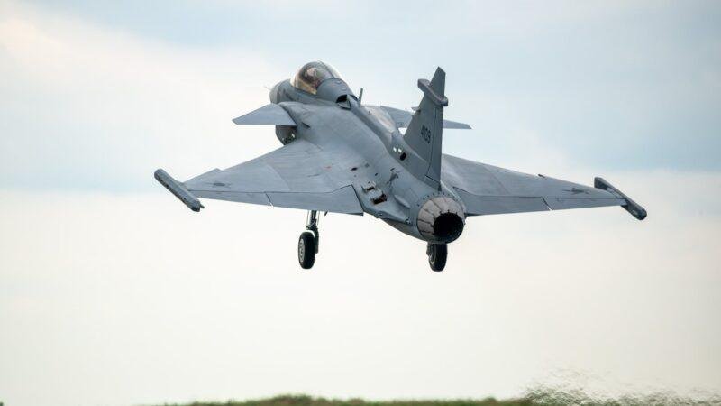 Brazil-built Gripen E fighter jet flying with landing gear extended during a low-altitude flight near an airfield.