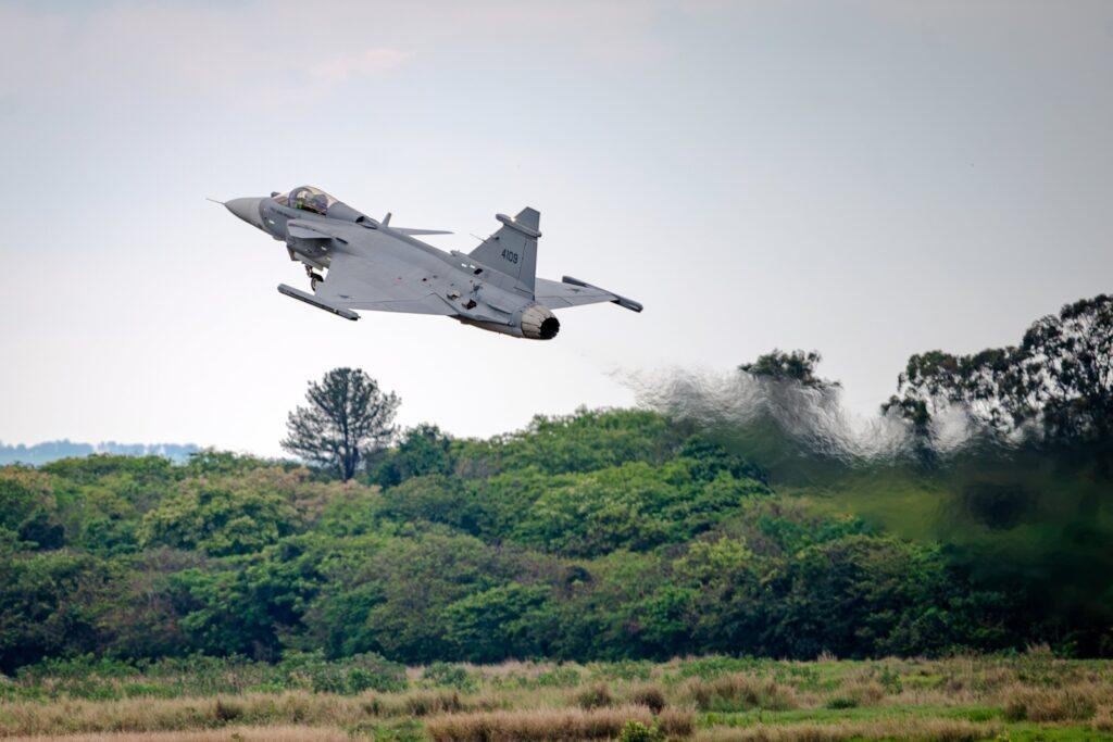 Brazil-built Gripen E fighter aircraft flying above green vegetation with its landing gear extended during an acceptance flight.