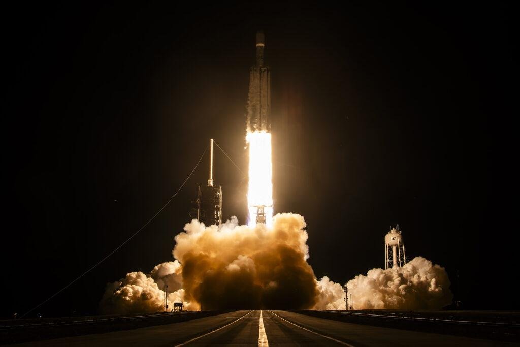 Falcon Heavy rocket lifting off at night on the NROL-97 mission, with a bright exhaust plume and clouds of smoke over Launch Complex 39A