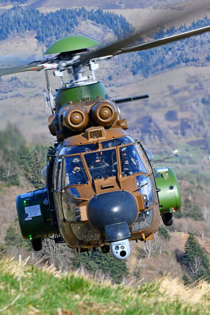 Airbus H215M military helicopter flying over a mountainous landscape with its cockpit and twin-engine configuration visible.