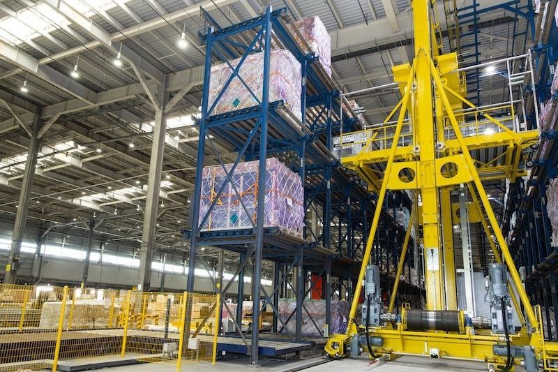 Automated air cargo warehouse with high-bay storage racks, palletized freight and a yellow mechanical lifting system.