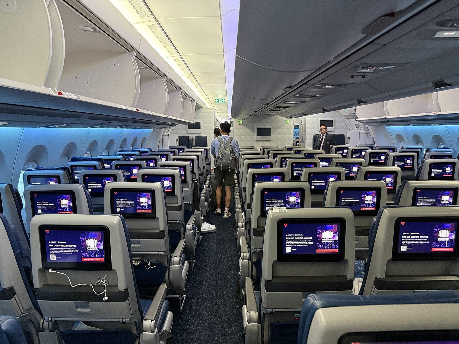 Widebody economy cabin with rows of Delta-branded seatback screens, overhead bins open and a passenger seen from behind walking down the aisle