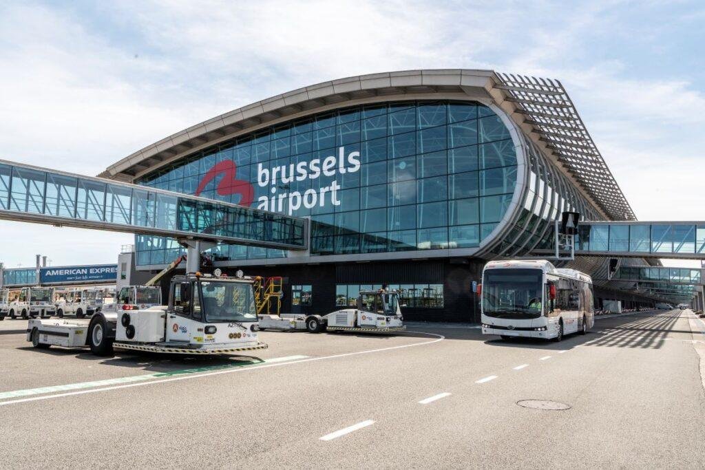 Brussels Airport strike article illustrated by the airport's glass-fronted passenger terminal, ground service vehicles, and airport shuttle bus.