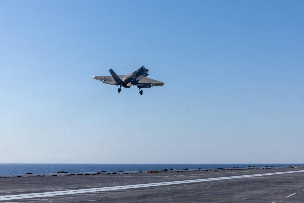 U.S. Navy F-35C Lightning II fighter airborne above an aircraft carrier flight deck with landing gear extended.