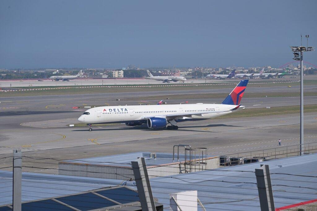 Delta Air Lines Airbus A350 in white livery with red and blue tail taxiing on an airport taxiway, with other airliners parked in the distance
