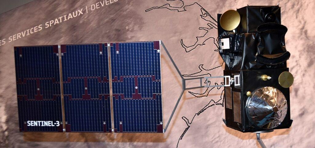 Scale model of the Sentinel-3 satellite with a three-panel solar array labelled SENTINEL-3, shown against an exhibition wall printed with a map