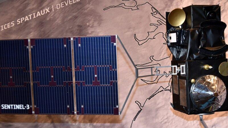 Scale model of the Sentinel-3 satellite with a three-panel solar array labelled SENTINEL-3, shown against an exhibition wall printed with a map