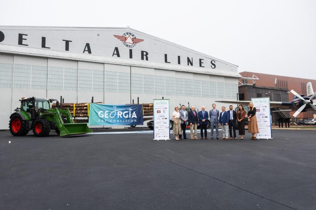 Nine people stand between Georgia SAF Coalition banners in front of the Delta Air Lines hangar, with a green tractor and a truck loaded with logs beside them
