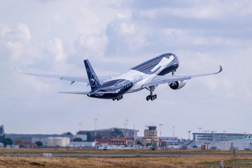 Lufthansa A350-1000 in special blue 100th-anniversary livery flying above an airport with landing gear extended.