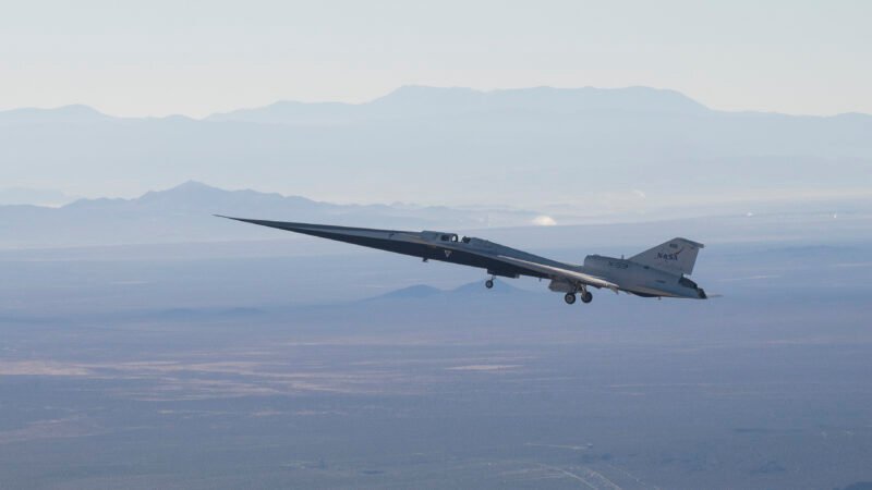 NASA’s X-59 flies above the Mojave Desert against a blue sky. The aircraft’s long nose is angled slightly upward to the left, with its landing gear down. Layers of mountains are visible in the background. The aircraft is backlit, appearing to float as it ferries to NASA’s Armstrong Flight Research Center in Edwards, California.