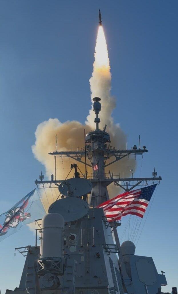 Missile climbing on a bright exhaust plume above the mast of a Navy destroyer flying the American flag
